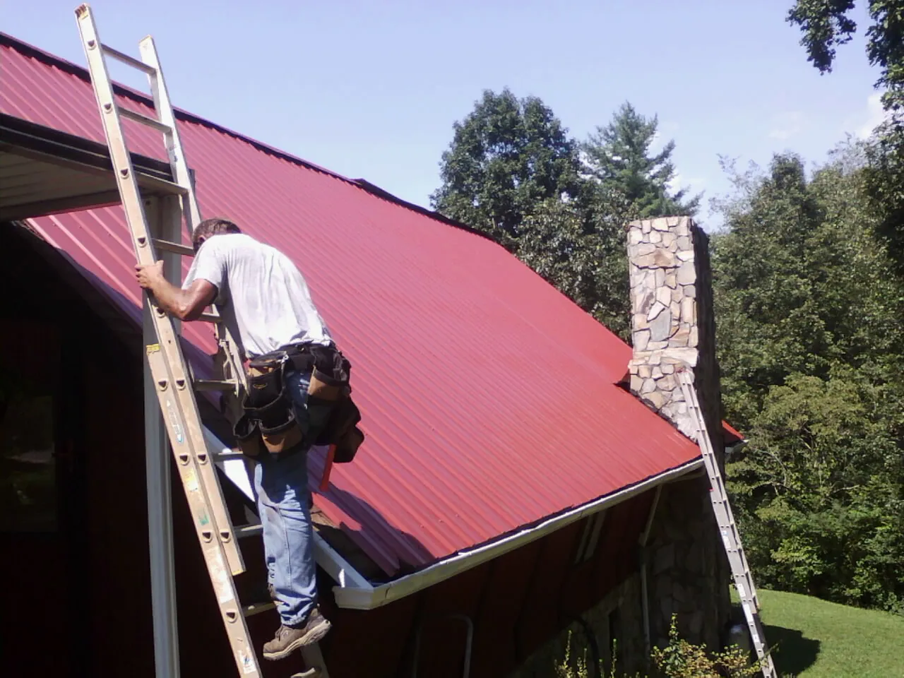Roofer on a ladder beside a red metal roof and stone chimney — Soffit & Fascia Repair by Roofing Brothers in Coats